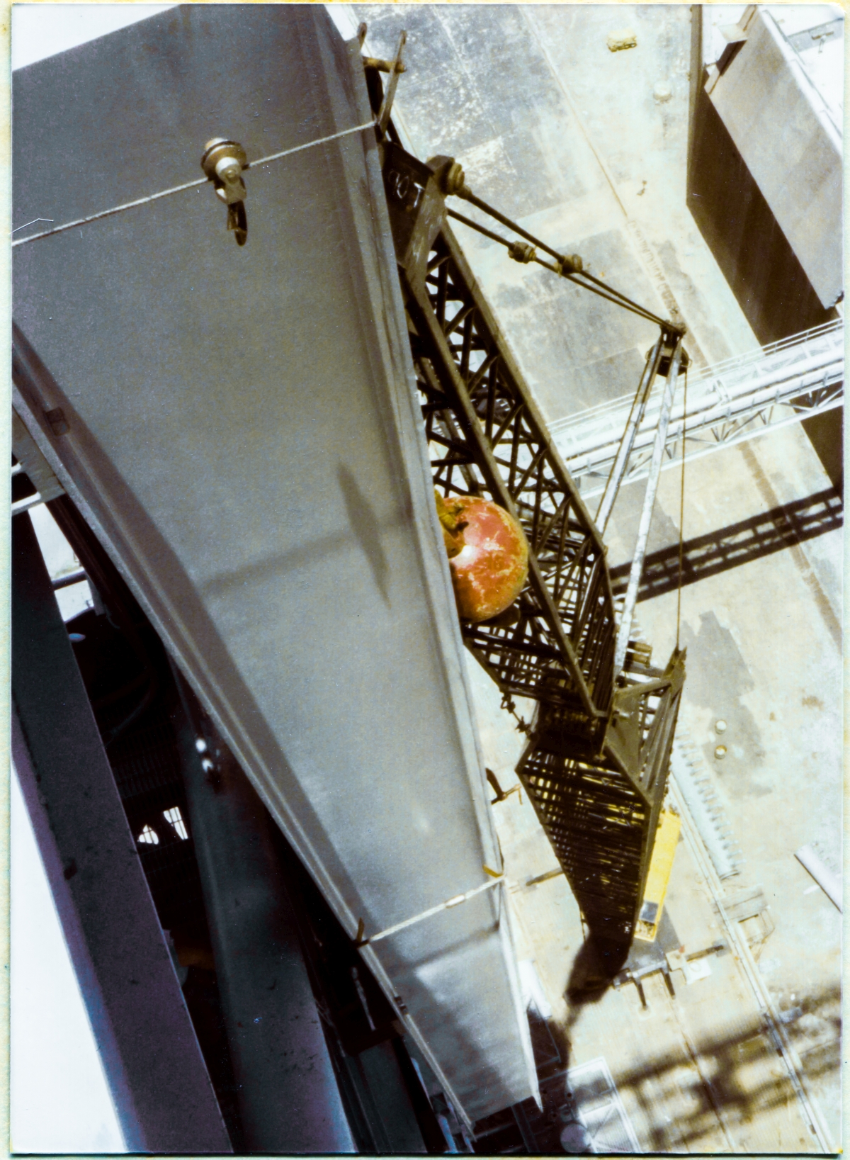 Image 081. You are on top of the Fixed Service Structure, 300 feet above the bottom of the Flame Trench at Space Shuttle Launch Complex 39-B, Kennedy Space Center, Florida, on the boom of the Hammerhead Crane, which projects outward to the east of the main body of the FSS. You are looking down the length of the web of one of the two 40-foot-long W24x104's which make up the main body of the GOX Arm Hinges Support Strongback, which is in suspension, held up by the yellow crane which you see a small part of, including the operator through his cab window, far beneath you sitting on the Pad Deck at the bottom end of a black boom which is almost 250 feet tall. High-summer midday shadows in Florida point nearly straight down beneath that which is casting them on elements of the world below. In the top right corner of the frame, the East Side Flame Deflector appears as a gray rectangle above the fifty-foot-high darkness of the East Flame Trench Wall. Immediately below it, the pale steel of the North Piping Bridge spans the Flame Trench, casting its latticework shadow on the refractory bricks that line its bottom surface. In the bottom right of the frame, the softer latticework shadow of the Hammerhead Crane, much farther above, which you are standing on, extends across the Crawlerway surface of the Pad Deck, to and beyond the invisible five-story precipice of the West Wall of the Flame Trench which is betrayed only by the very slight offset in the shadow, near the right margin of the frame. Your own shadow is in-frame, but unseeable, dwarfed into invisibility by the imagination-beggaring scale of that which you are a part of. You are, if only temporarily, part of an Ironworker's World, and it is a world apart, like no other. Anywhere. Photo by James MacLaren.